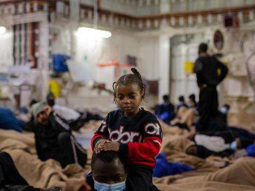 A child stands with a caregiver on the deck of a search and rescue ship assisting refugees and asylum seekers.