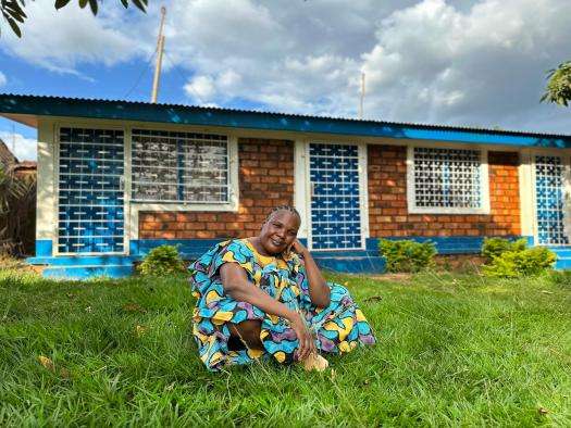 A smiling woman in brightly colored dress sits on the lawn in front of a brick building in Central African Republic.