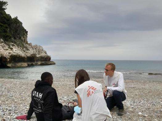 A man sits on the shores of Greece and is tended to by two MSF workers. 