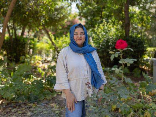 A woman wearing a blue headscarf stands next to a red rose in Greece. 
