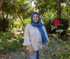 A woman wearing a blue headscarf stands next to a red rose in Greece. 