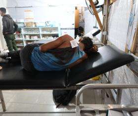 A teenage boy with a bandaged and bloody shoulder lies on a hospital bed in Gaza, Palestine. 
