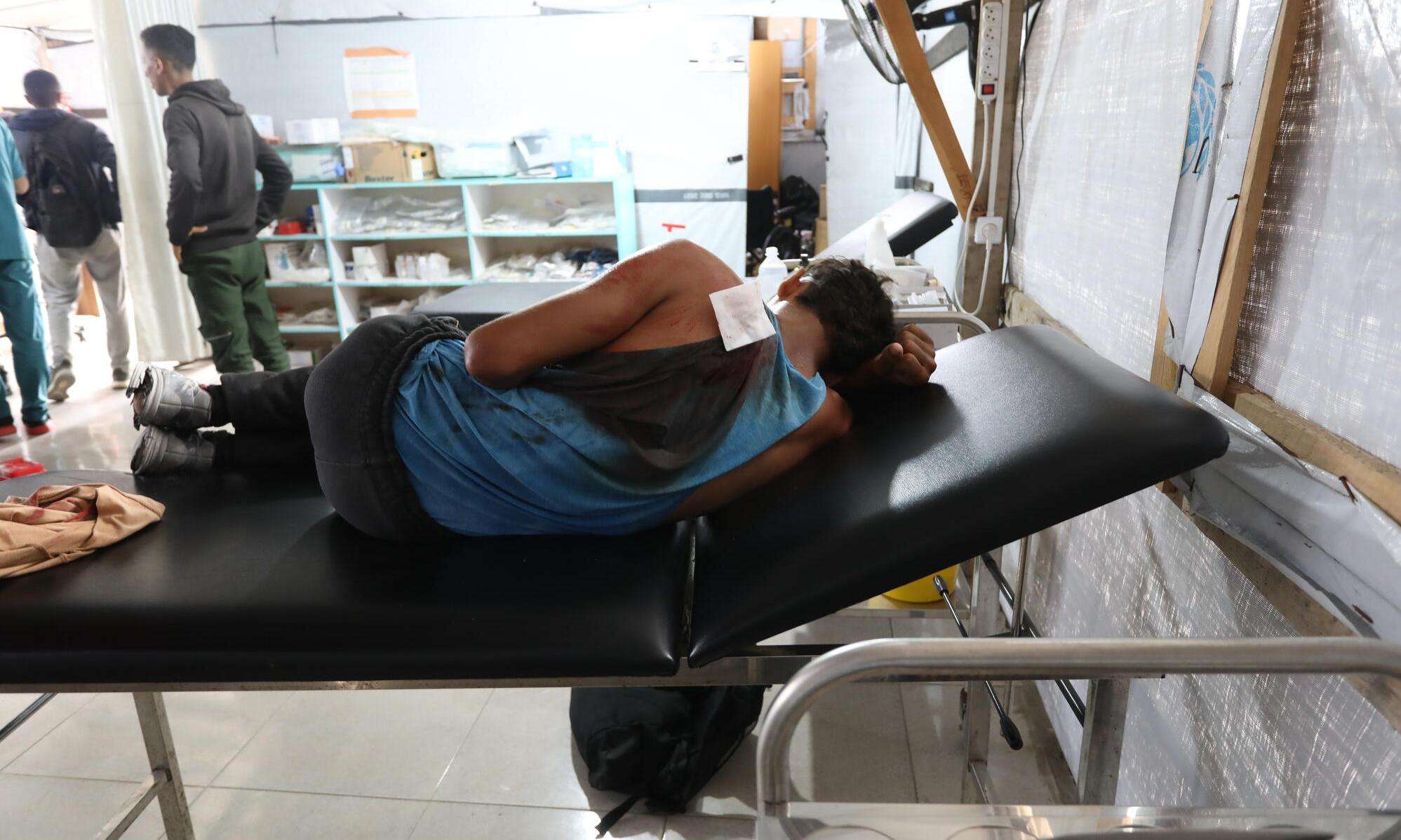 A teenage boy with a bandaged and bloody shoulder lies on a hospital bed in Gaza, Palestine. 