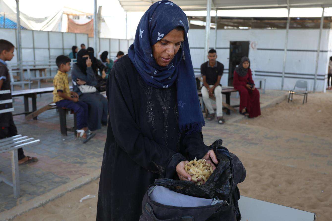 A woman stands next to a black plastic bag filled with pasta in Gaza, Palestine.
